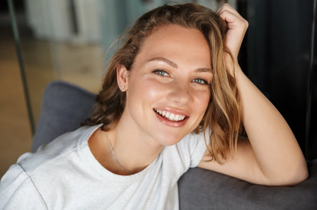 Young woman smiling brightly while touching her hair, wearing white t-shirt in indoor setting.