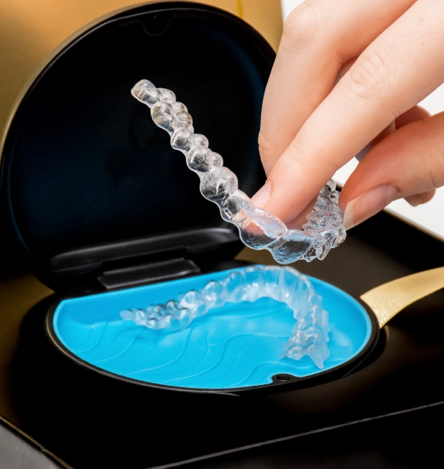 Hand holding clear dental aligner over blue tray with black case in background.