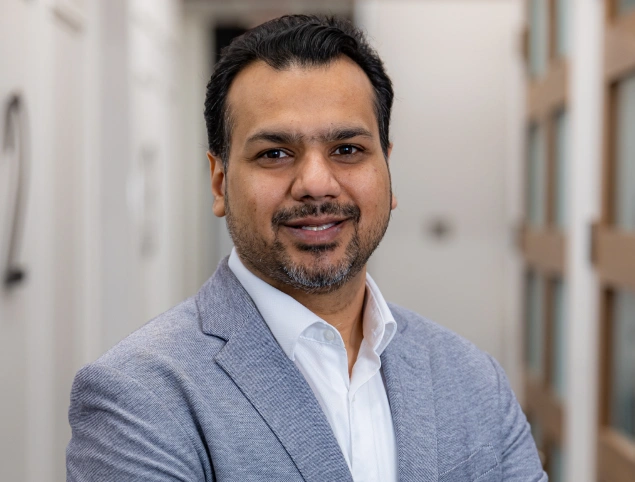 Professional male dentist in blue blazer smiling at camera in modern dental office hallway.